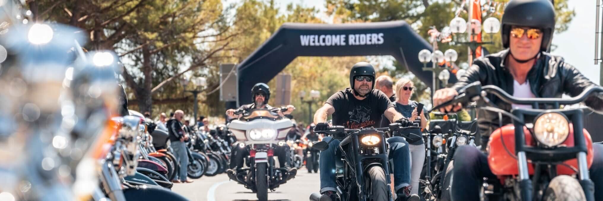 A group of motorcyclists riding through a parade-style street event with a large black arch that reads 'WELCOME RIDERS.' The scene is outdoors with trees and blurred motorcycles in the foreground and background.